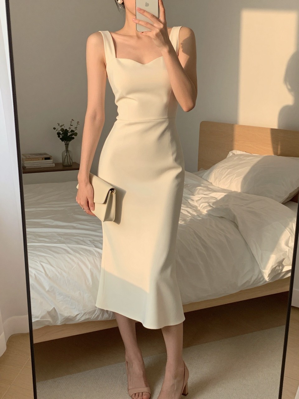mirror selfie of a woman wearing a buttercream crepe midi dress, pearl stud earrings, nude block heels, and a small cream clutch, holding a phone in front of a mirror, cropped at NECK so face doesn't show, with a natural casual selfie pose, in a feminine-themed bedroom, evening golden hour, including small lived-in details like soft bedding in the background, latest iPhone photo quality.