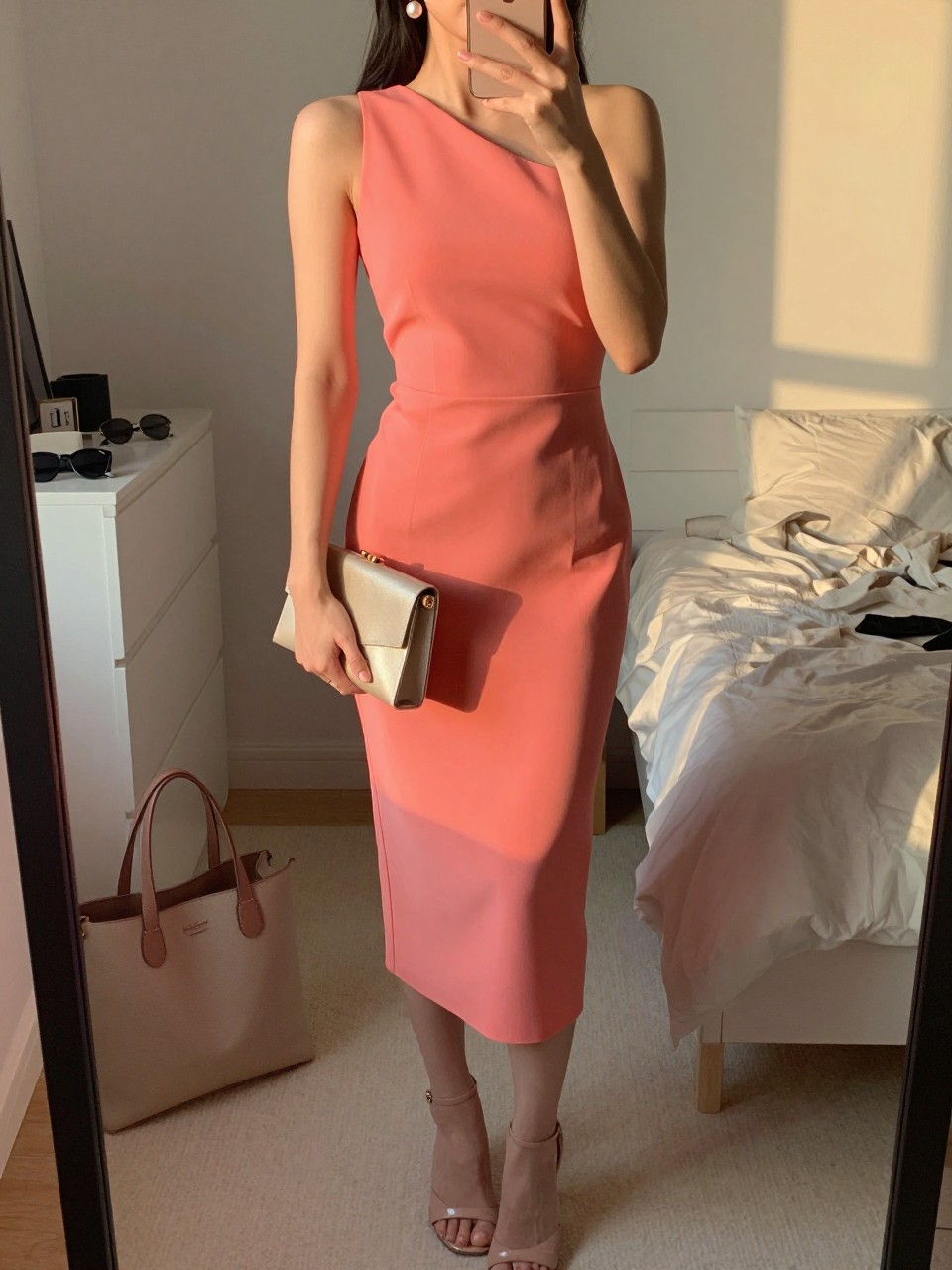 mirror selfie of a woman wearing a coral one-shoulder crepe dress, pearl drop earrings, nude strappy heels, and a small structured neutral clutch, holding a phone in front of a mirror, cropped at NECK so face doesn't show, with a natural casual selfie pose, in a feminine-themed bedroom, evening golden hour, including small lived-in details like a tote bag on the floor, sunglasses on a dresser, or soft bedding in the background, latest iPhone photo quality.