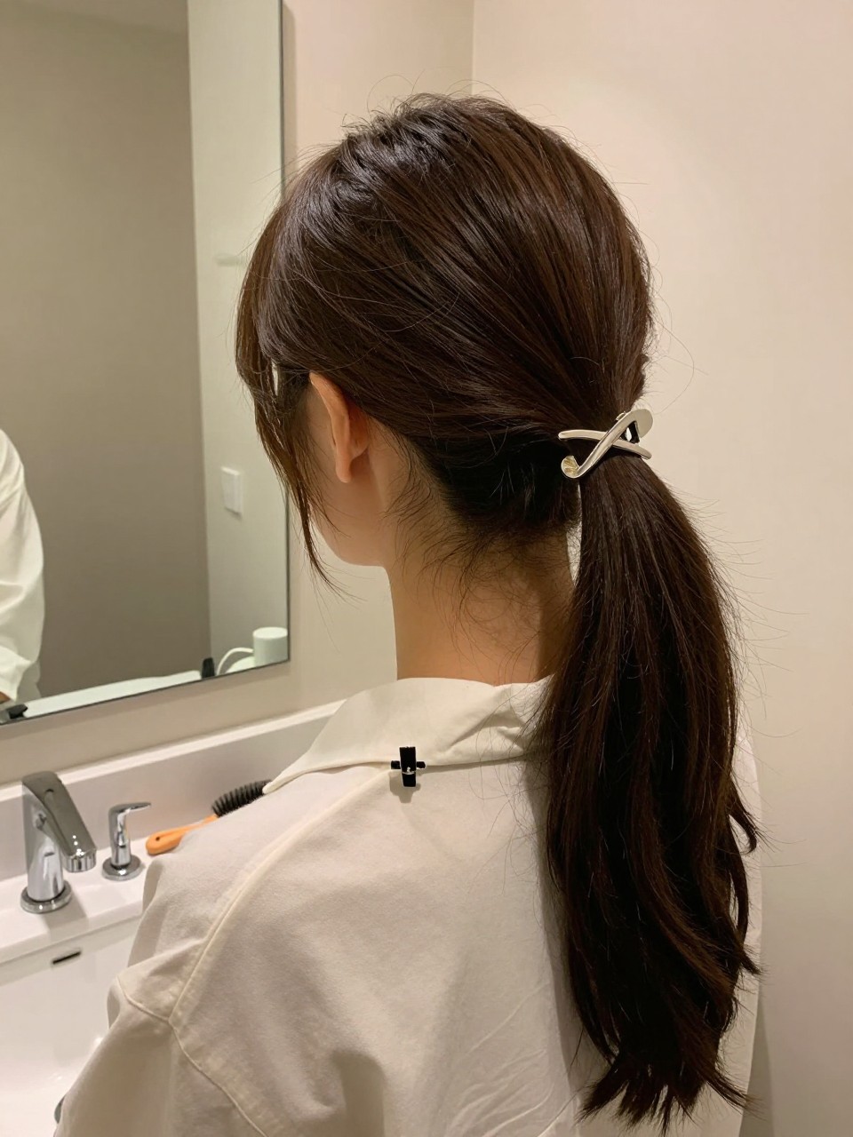 Photo of a woman wearing a sleek low ponytail with a statement barrette, back-of-head view to highlight the barrette detail, setting in a bathroom vanity mirror, lighting soft indoor lighting, containing a small lived-in detail such as a hair clip clipped to the collar and a hairbrush on the vanity, latest iPhone photo quality.