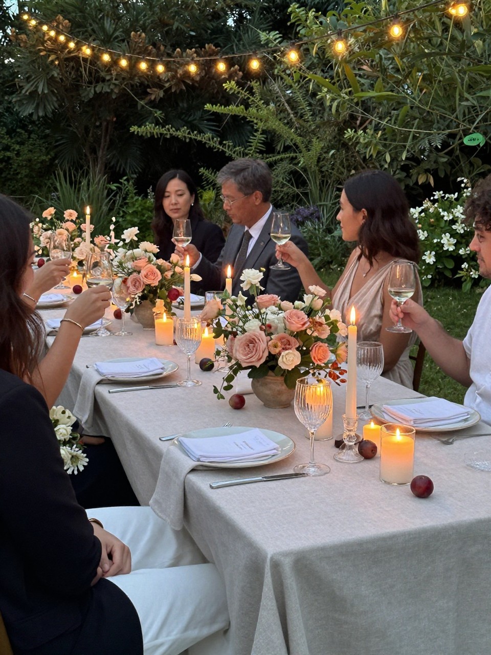 Photo of a late summer garden reception table under string lights with neutral linens, dusty peach florals, small plum accents, and candles glowing, camera angle: over-the-shoulder candid, setting: outdoor garden reception tables, lighting: candlelight glow, containing subtle real wedding activity such as guests clinking glasses and a bouquet resting on a chair nearby, latest iPhone photo quality.