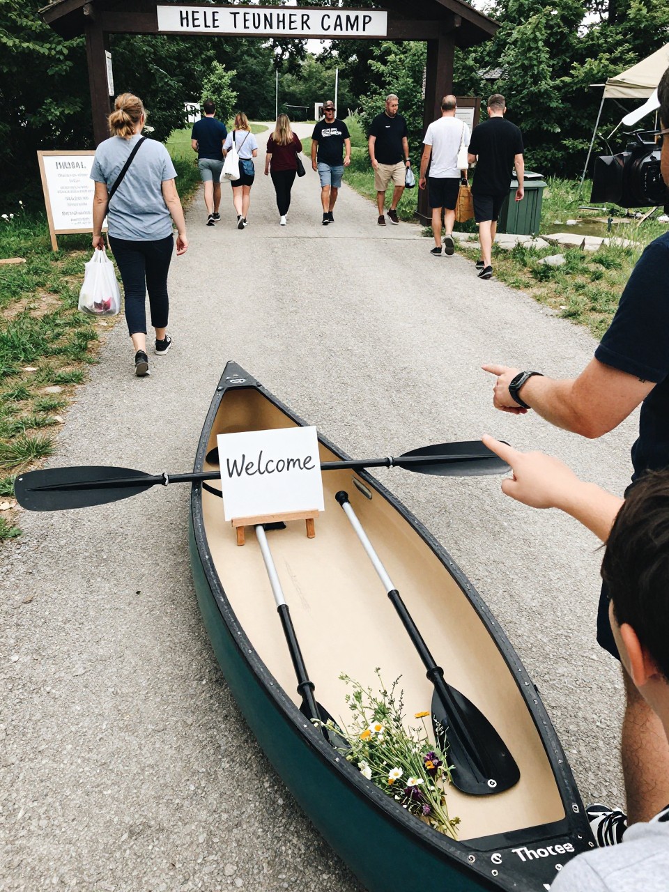 Photo of a canoe parked near a summer camp entrance with a welcome sign resting inside, a couple paddles, and small wildflower bundles, camera angle: over-the-shoulder candid, setting: camp check-in area by the main path, lighting: natural daylight, containing subtle real wedding activity such as guests walking up with weekend bags and someone pointing at the sign, latest iPhone photo quality.