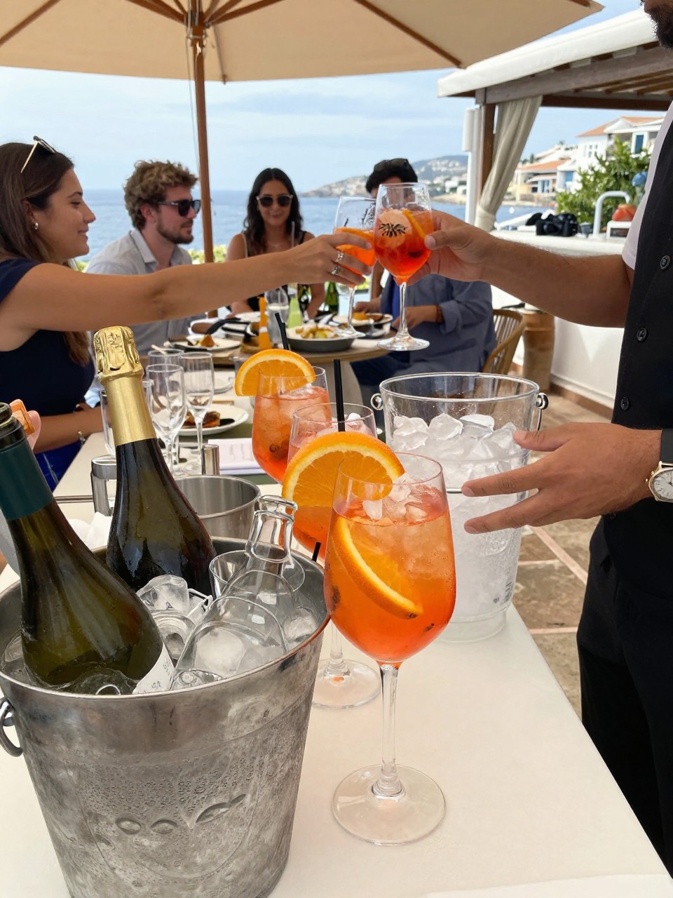 Photo of an aperitivo spritz welcome station on a Mediterranean terrace with bottles, ice buckets, sliced oranges, and glasses ready, camera angle: detail close-up, setting: terrace cocktail hour area, lighting: natural daylight, containing subtle real wedding activity such as guests reaching for drinks and a server topping up ice, latest iPhone photo quality.