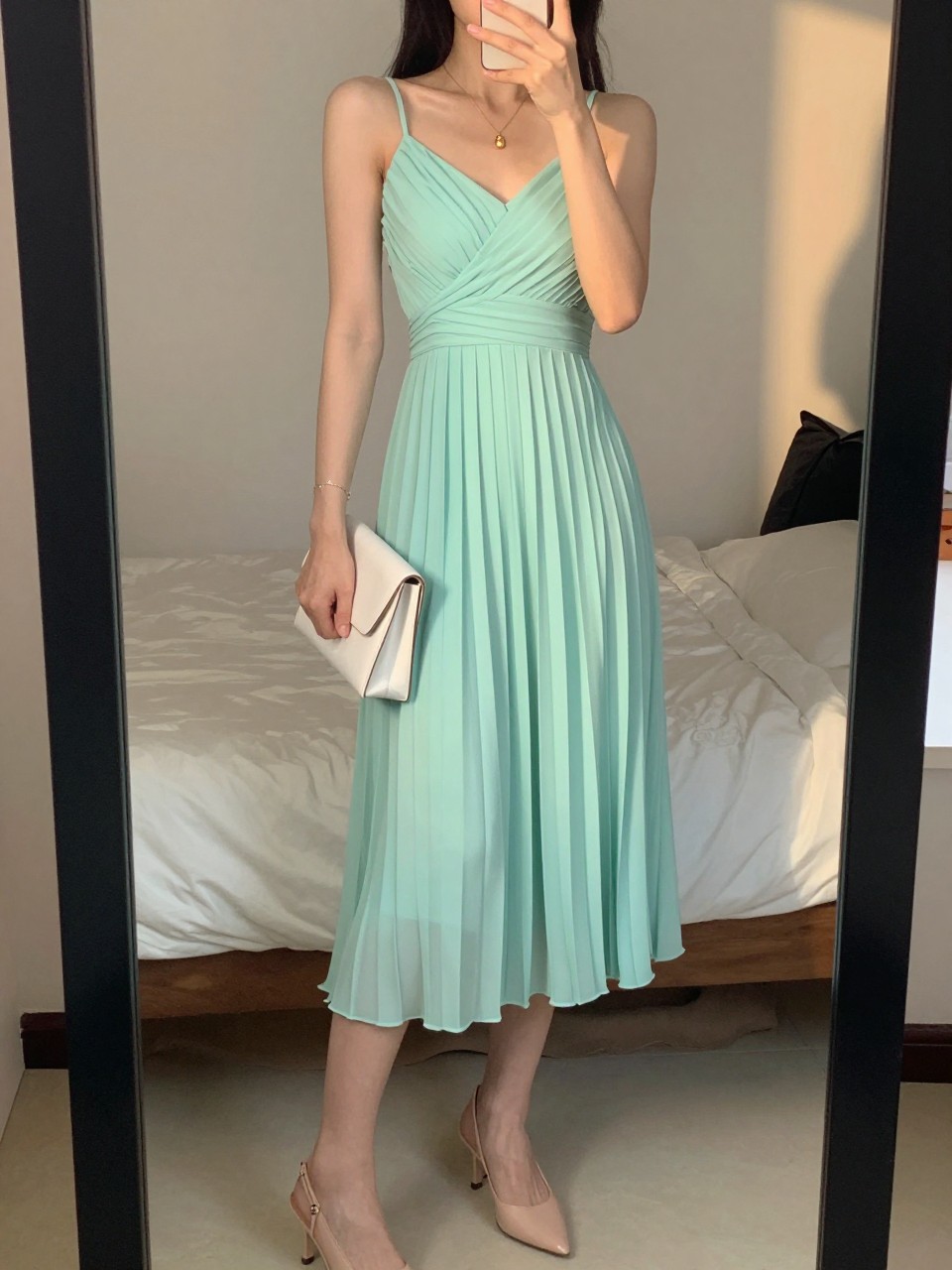 mirror selfie of a woman wearing a mint pleated chiffon midi dress, nude slingback heels, a delicate gold pendant necklace, and a small white clutch, holding a phone in front of a mirror, cropped at NECK so face doesn't show, with a natural casual selfie pose, in a feminine-themed bedroom, evening golden hour, including small lived-in details like a soft bedding in the background, latest iPhone photo quality.