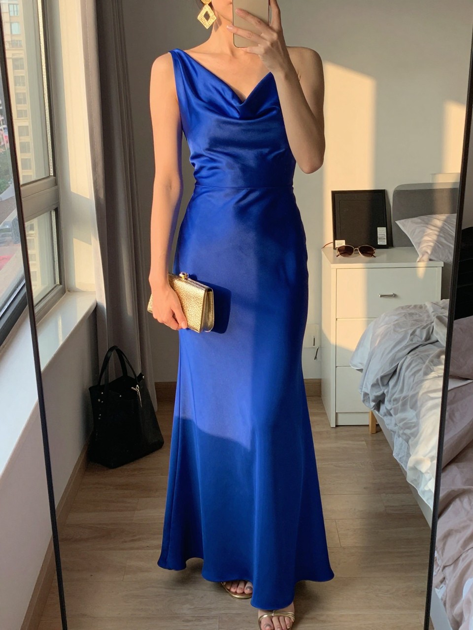 mirror selfie of a woman wearing a cobalt blue floor-length satin gown with a draped neckline, gold statement drop earrings, gold strappy heels, and a small gold clutch, holding a phone in front of a mirror, cropped at NECK so face doesn't show, with a natural casual selfie pose, in a city apartment, evening golden hour, including small lived-in details like a tote bag on the floor, sunglasses on a dresser, or soft bedding in the background, latest iPhone photo quality.