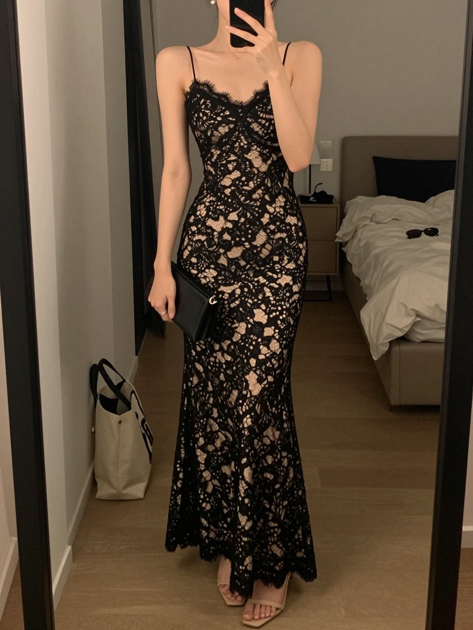 mirror selfie of a woman wearing a fitted black lace floor-length gown with a smooth nude slip lining, nude strappy heels, small crystal stud earrings, and a black clutch with a subtle shine, holding a phone in front of a mirror, cropped at NECK so face doesn't show, with a natural casual selfie pose, in a city apartment, warm moody lighting, including small lived-in details like a tote bag on the floor, sunglasses on a dresser, or soft bedding in the background, latest iPhone photo quality.
