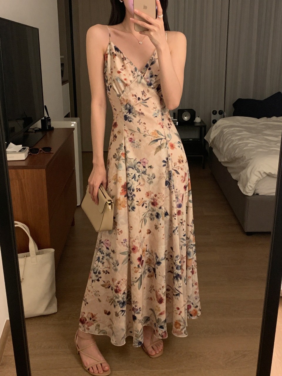 mirror selfie of a woman wearing a soft floral printed maxi dress with a flowy skirt, nude strappy sandals, small crystal stud earrings, and a small neutral clutch, holding a phone in front of a mirror, cropped at NECK so face doesn't show, with a natural casual selfie pose, in a city apartment, warm moody lighting, including small lived-in details like a tote bag on the floor, sunglasses on a dresser, or soft bedding in the background, latest iPhone photo quality.