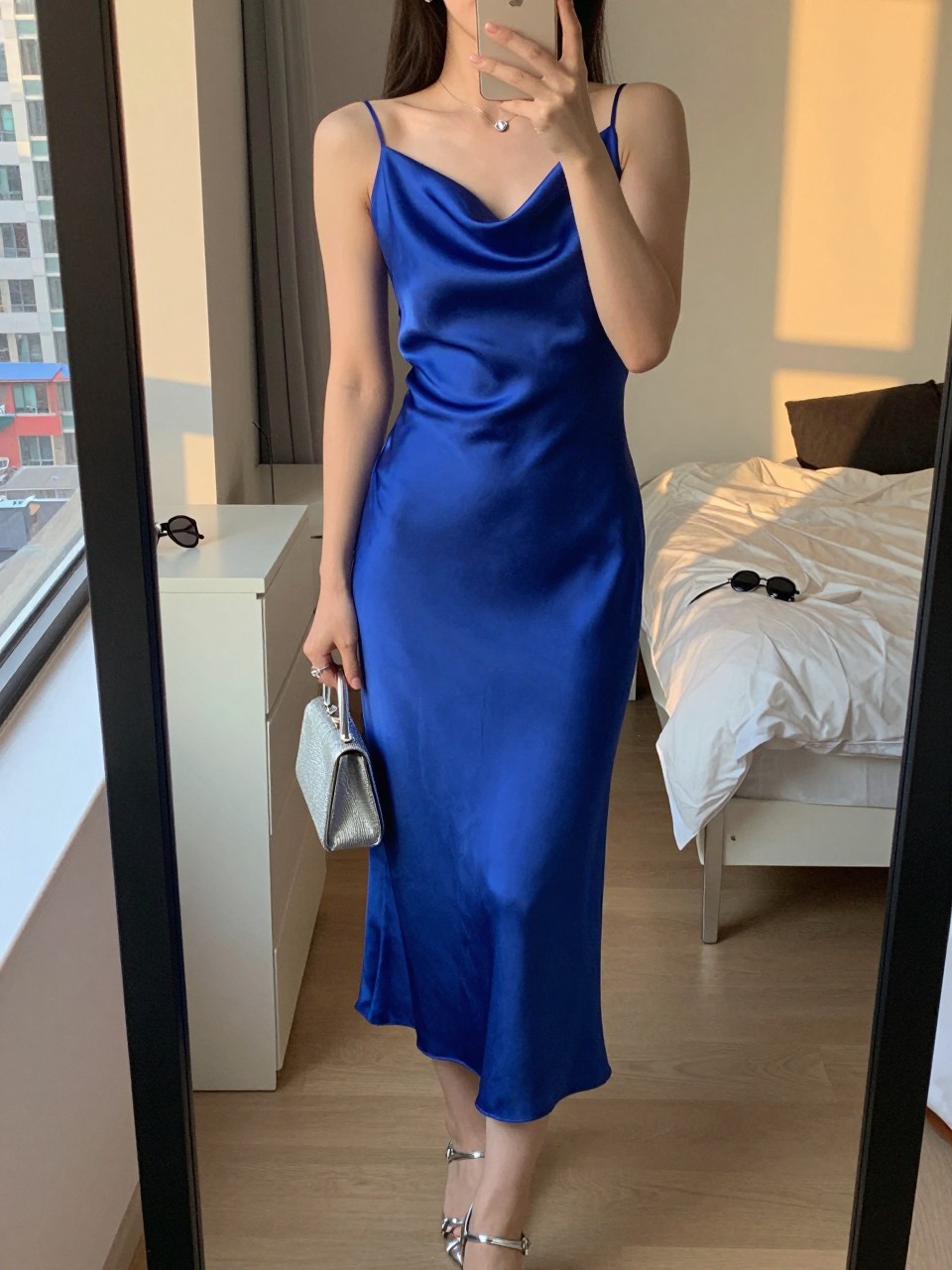 mirror selfie of a woman wearing a cobalt satin midi dress with a draped neckline, silver strappy heels, silver stud earrings, and a small silver clutch, holding a phone in front of a mirror, cropped at NECK so face doesn't show, with a natural casual selfie pose, in a city apartment, evening golden hour, including small lived-in details like a, sunglasses on a dresser, or soft bedding in the background, latest iPhone photo quality.