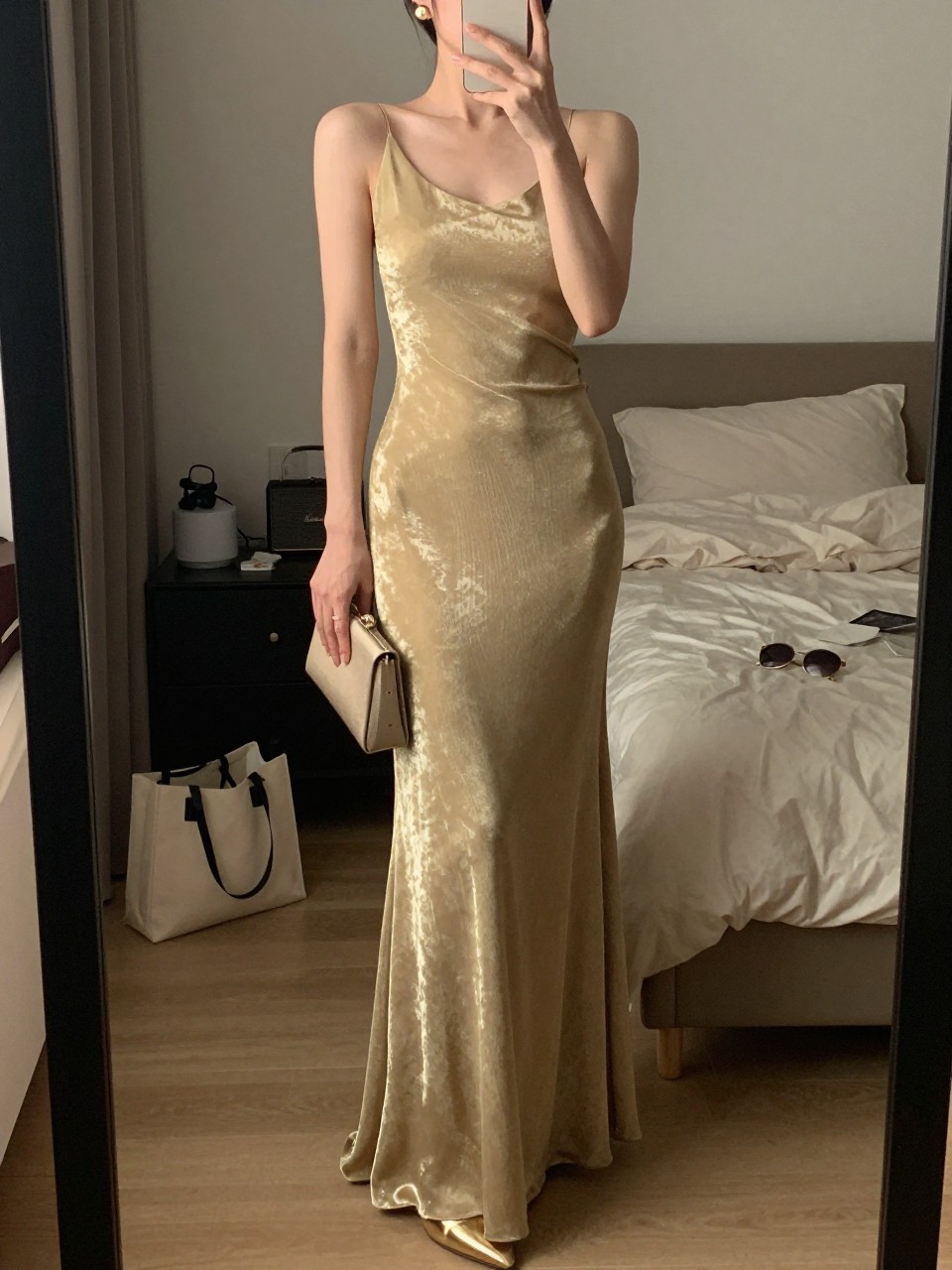 mirror selfie of a woman wearing a floor-length gold metallic gown with a clean silhouette, minimal gold heels, simple gold stud earrings, and a small neutral clutch, hair in a sleek low bun, holding a phone in front of a mirror, cropped at NECK so face doesn't show, with a natural casual selfie pose, in a feminine-themed bedroom, warm moody lighting, including small lived-in details like a tote bag on the floor, sunglasses on a dresser, or soft bedding in the background, latest iPhone photo quality.