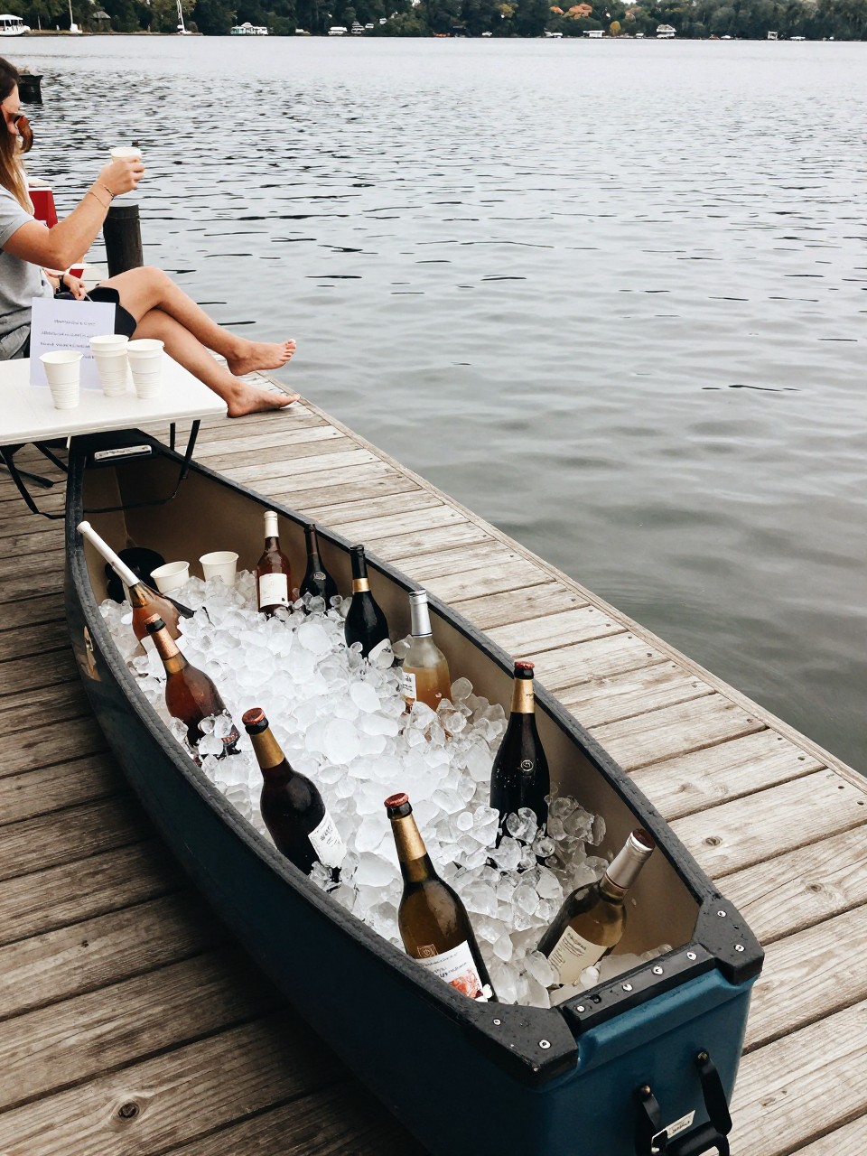 Photo of a canoe filled with ice and bottled drinks set up as a cooler near a lakeside dock with cups and a simple sign, camera angle: guest table angle, setting: lakeside dock hangout area, lighting: natural daylight, containing subtle real wedding activity such as guests grabbing drinks and sitting on the dock with feet near the water, latest iPhone photo quality.