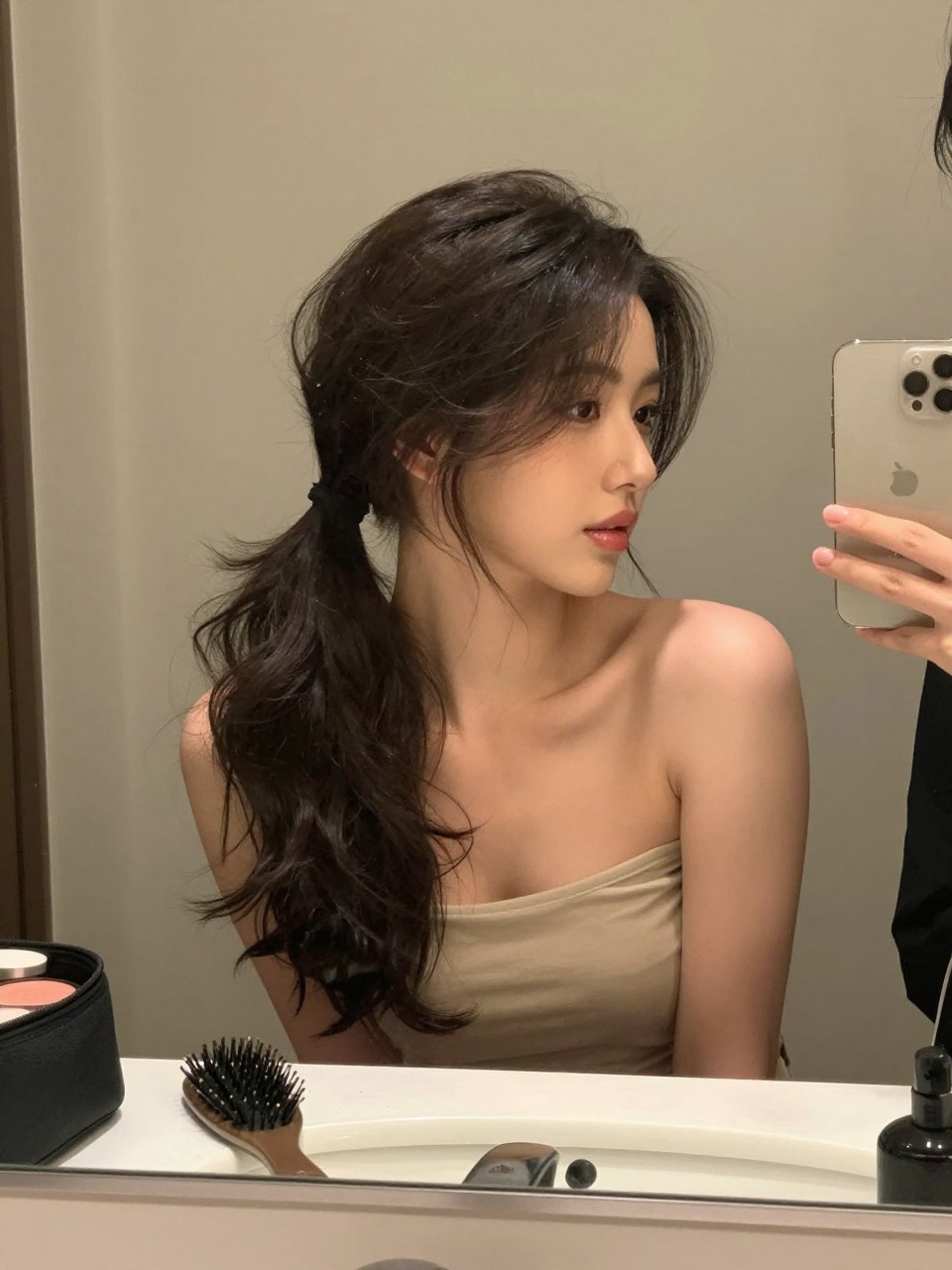Photo of a woman wearing a low ponytail with soft face-framing waves, candid mirror selfie, setting in a bathroom vanity mirror, lighting soft indoor lighting, containing a small lived-in detail such as a hairbrush on the vanity and a makeup bag nearby, latest iPhone photo quality.