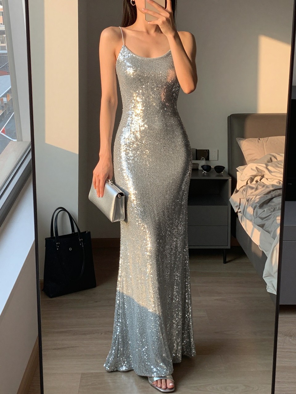 mirror selfie of a woman wearing a floor-length silver sequin gown with a simple silhouette, thin-strap silver heels, small silver hoop earrings, and a smooth silver clutch, holding a phone in front of a mirror, cropped at NECK so face doesn't show, with a natural casual selfie pose, in a city apartment, evening golden hour, including small lived-in details like a tote bag on the floor, sunglasses on a dresser, or soft bedding in the background, latest iPhone photo quality.