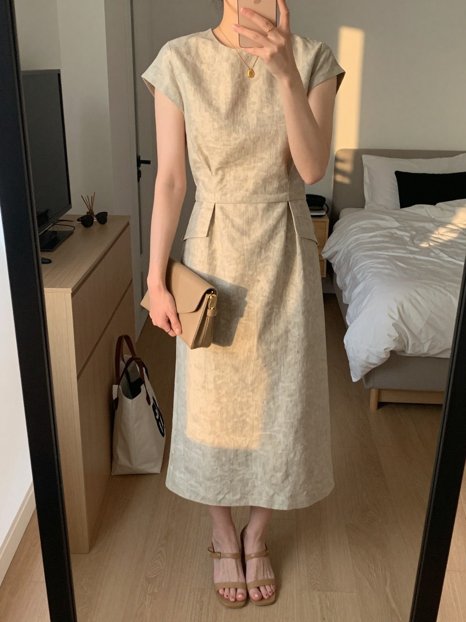 mirror selfie of a woman wearing a sand linen blend midi dress with a tailored waist, tan heeled sandals, a delicate gold pendant necklace, and a small tan clutch, holding a phone in front of a mirror, cropped at NECK so face doesn't show, with a natural casual selfie pose, in a city apartment, evening golden hour, including small lived-in details like a tote bag on the floor, sunglasses on a dresser, or soft bedding in the background, latest iPhone photo quality.