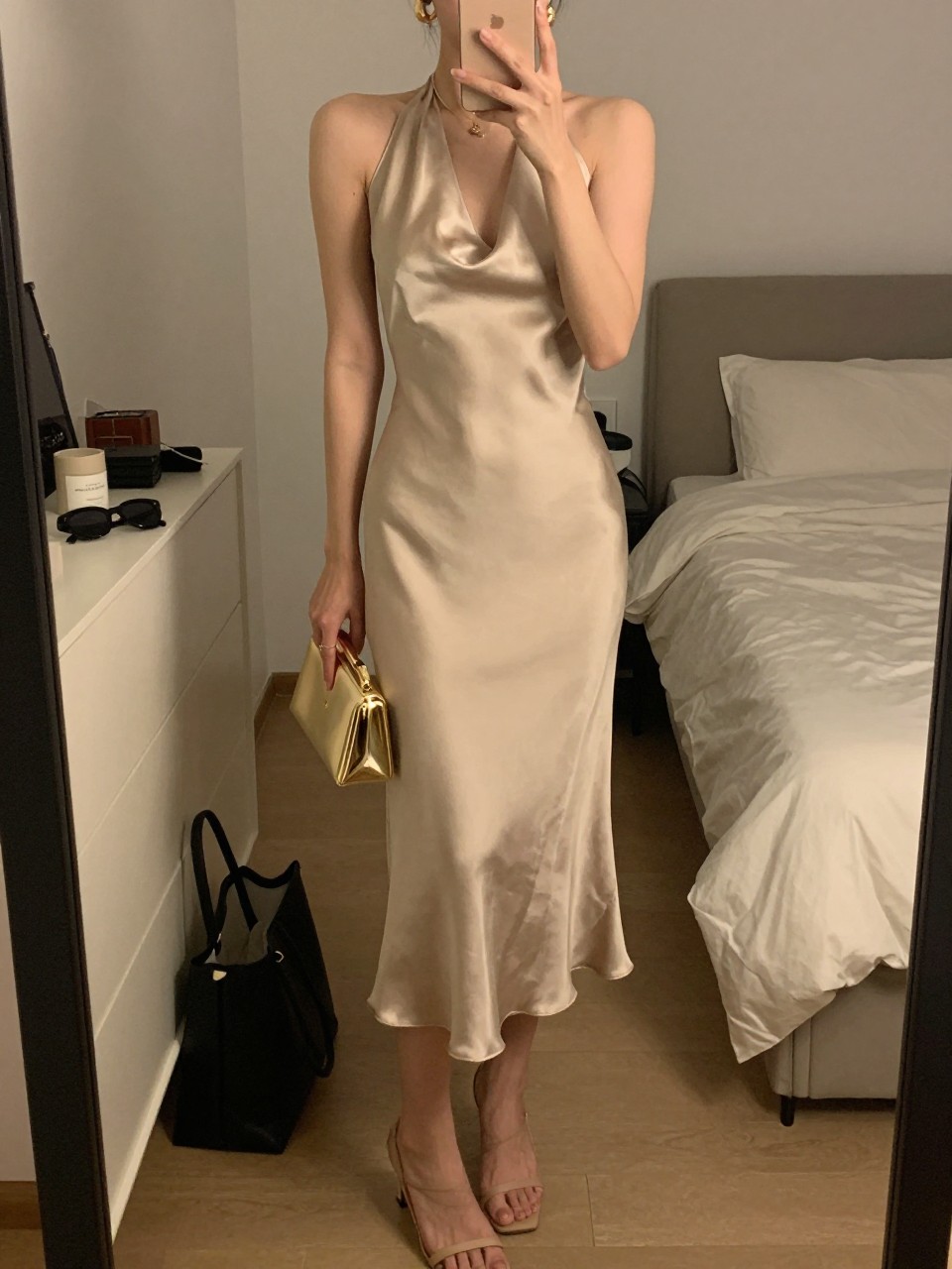 mirror selfie of a woman wearing a champagne satin halter midi dress, medium gold hoop earrings, nude strappy heels, and a small gold clutch, holding a phone in front of a mirror, cropped at NECK so face doesn't show, with a natural casual selfie pose, in a feminine-themed bedroom, warm moody lighting, including small lived-in details like a tote bag on the floor, sunglasses on a dresser, or soft bedding in the background, latest iPhone photo quality.