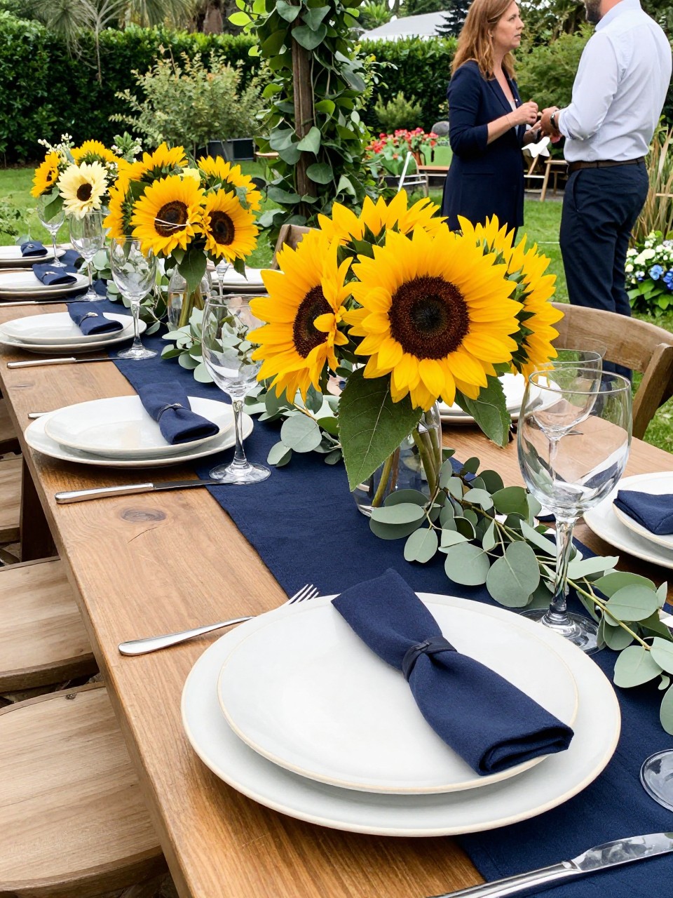 Photo of a late summer garden reception table with navy runners, sunflower arrangements, neutral plates, and soft greenery, camera angle: detail close-up, setting: outdoor garden venue reception, lighting: natural daylight, containing subtle real wedding activity such as guests chatting and a bouquet resting nearby, latest iPhone photo quality.