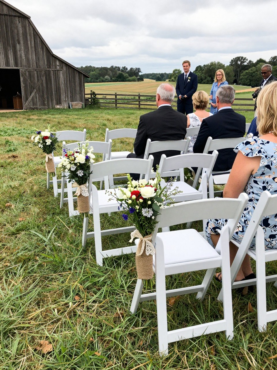 Photo of an outdoor countryside ceremony aisle with chairs lined up in a grassy field and burlap and lace wildflower bundles tied to the aisle chairs, camera angle: wide ceremony view, setting: countryside field ceremony space near a barn, lighting: natural daylight, containing subtle real wedding activity such as guests settling into seats and a bouquet resting on the front row chair, latest iPhone photo quality.