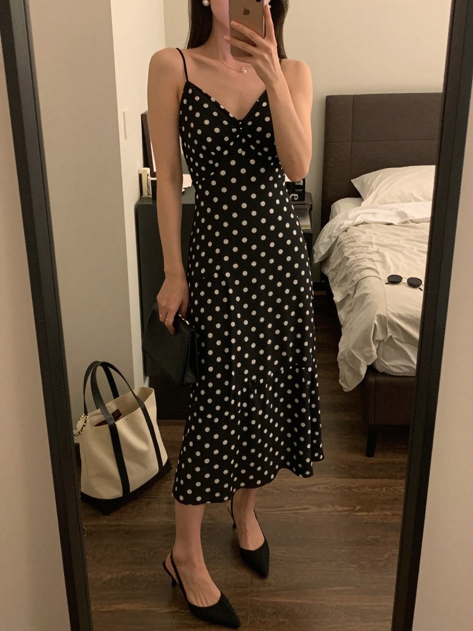 mirror selfie of a woman wearing a black and white polka dot chiffon midi dress, black slingback heels, pearl stud earrings, and a small black clutch, holding a phone in front of a mirror, cropped at NECK so face doesn't show, with a natural casual selfie pose, in a city apartment, warm moody lighting, including small lived-in details like a tote bag on the floor, sunglasses on a dresser, or soft bedding in the background, latest iPhone photo quality.