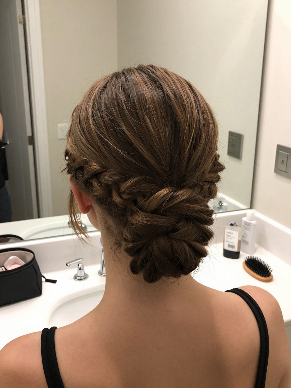 Photo of a woman wearing a braided low bun with a clean center part, back-of-head view to highlight the braid wrap, setting in a bathroom vanity mirror, lighting soft indoor lighting, containing a small lived-in detail such as a makeup bag nearby and a hairbrush on the vanity, latest iPhone photo quality.