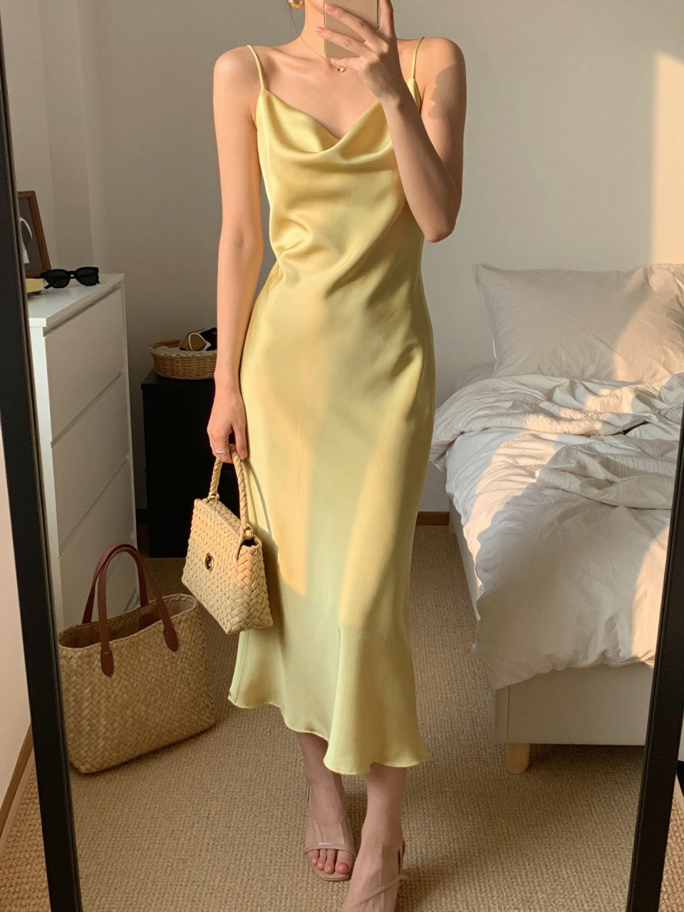 mirror selfie of a woman wearing a lemon yellow satin midi dress with a soft drape, nude strappy heels, small gold hoop earrings, and a structured woven mini bag, holding a phone in front of a mirror, cropped at NECK so face doesn't show, with a natural casual selfie pose, in a feminine-themed bedroom, evening golden hour, including small lived-in details like a tote bag on the floor, sunglasses on a dresser, or soft bedding in the background, latest iPhone photo quality.