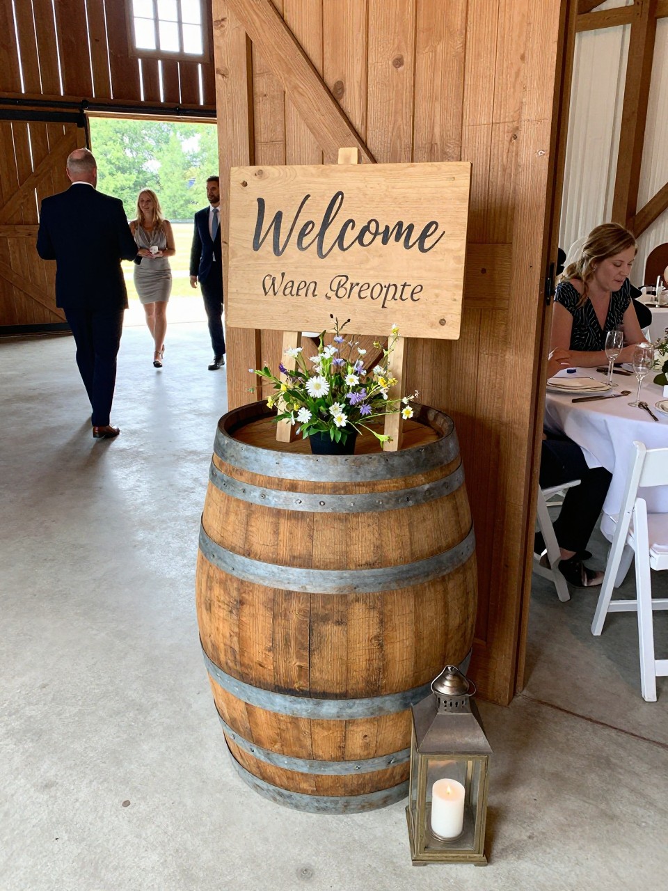 Photo of a wooden barrel welcome sign display near barn doors with a small wildflower arrangement and a lantern at the base, camera angle: guest table angle, setting: barn entrance welcome area, lighting: natural daylight, containing subtle real wedding activity such as guests walking in and someone pausing to read the sign, latest iPhone photo quality.