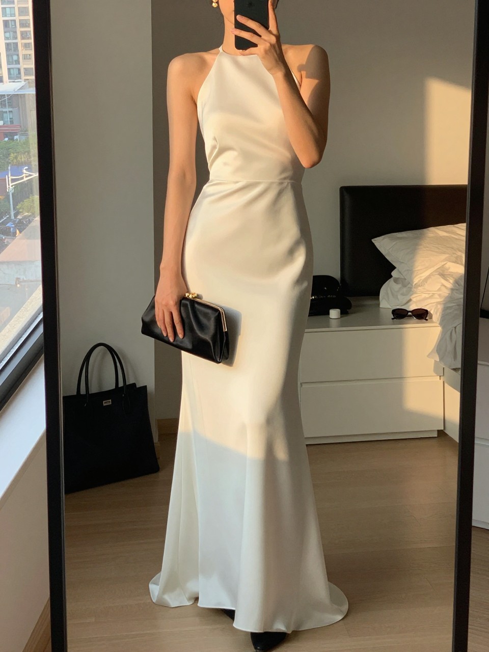 mirror selfie of a woman wearing an ivory halter floor-length gown in satin or structured crepe, minimal black heels, small gold drop earrings, and a black satin clutch, holding a phone in front of a mirror, cropped at NECK so face doesn't show, with a natural casual selfie pose, in a city apartment, evening golden hour, including small lived-in details like a tote bag on the floor, sunglasses on a dresser, or soft bedding in the background, latest iPhone photo quality.