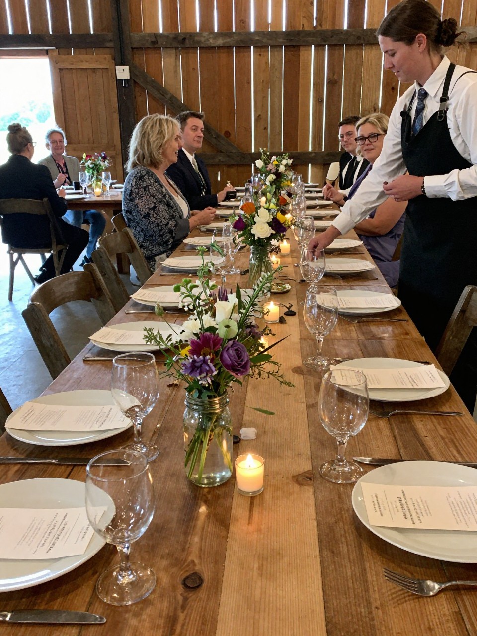Photo of rustic barn reception tables with wooden surfaces, mason jar wildflower centerpieces, and tea lights scattered between place settings, camera angle: guest table angle, setting: barn interior reception tables, lighting: soft reception lighting, containing subtle real wedding activity such as guests chatting and a server placing glasses on the table, latest iPhone photo quality.