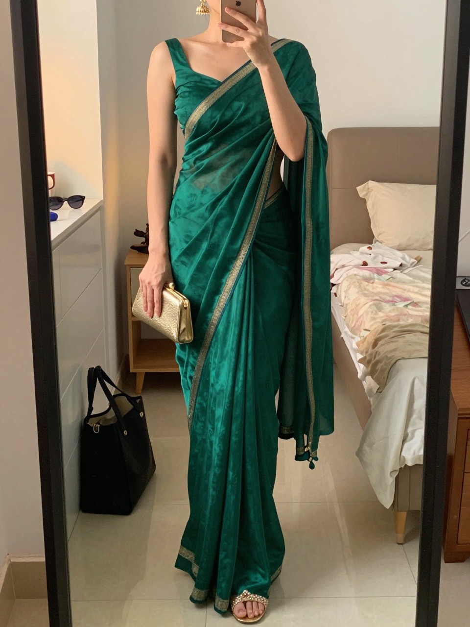 mirror selfie of a woman wearing an emerald silk saree with a subtle border and a fitted matching blouse, gold jhumka earrings, gold embellished heels, and a small gold clutch, holding a phone in front of a mirror, cropped at NECK so face doesn't show, with a natural casual selfie pose, in a feminine-themed bedroom, evening golden hour, including small lived-in details like a tote bag on the floor, sunglasses on a dresser, or soft bedding in the background, latest iPhone photo quality.