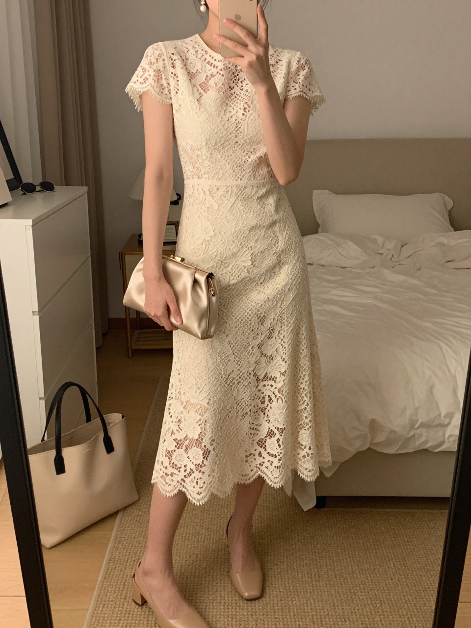 mirror selfie of a woman wearing a cream lace midi dress with a lined skirt, nude low block heels, pearl drop earrings, and a champagne satin clutch, holding a phone in front of a mirror, cropped at NECK so face doesn't show, with a natural casual selfie pose, in a feminine-themed bedroom, warm moody lighting, including small lived-in details like a tote bag on the floor, sunglasses on a dresser, or soft bedding in the background, latest iPhone photo quality.