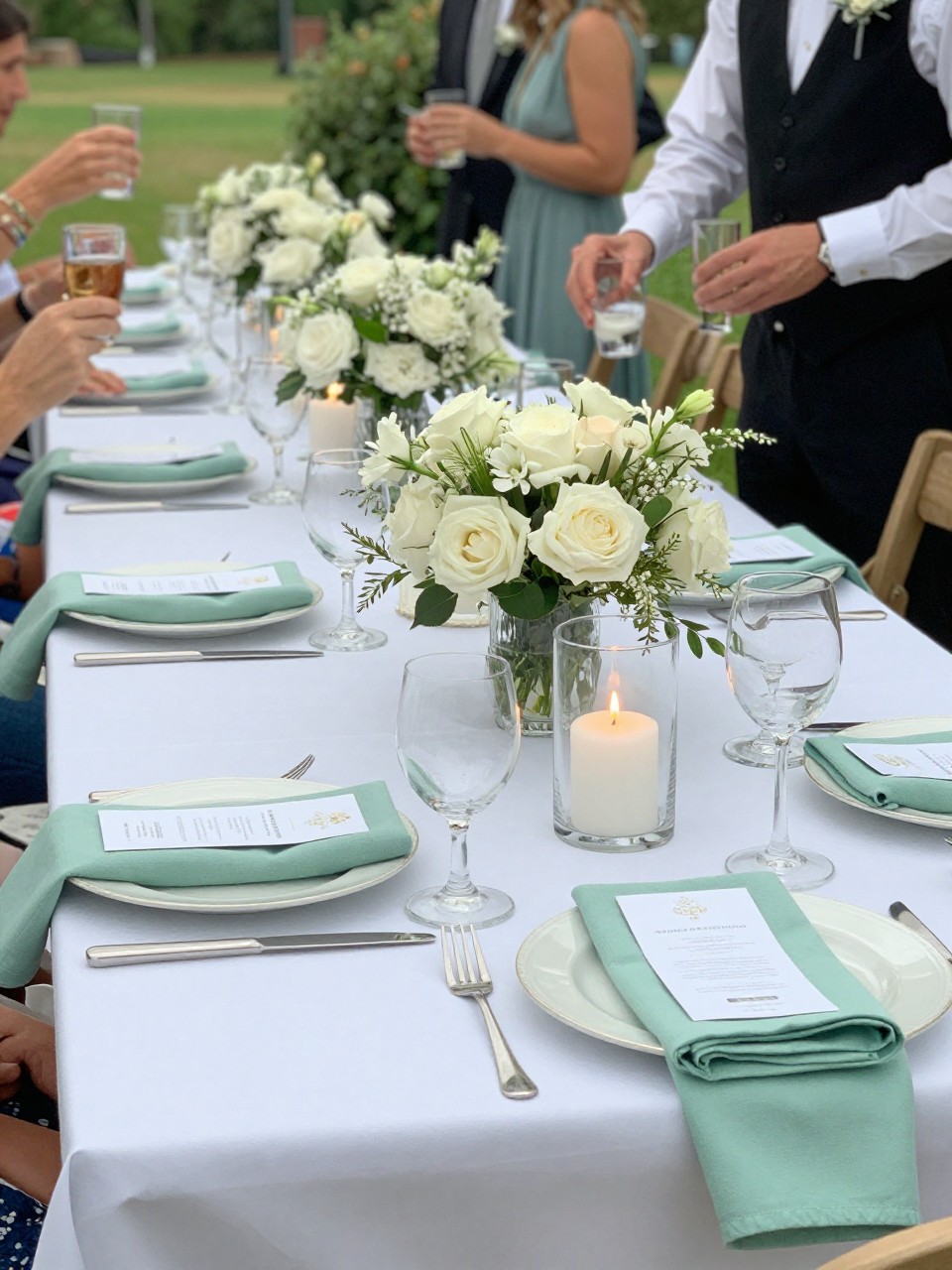 Photo of a late summer terrace reception table with pearl white linens, mint napkins, white florals, and clear glass candles, camera angle: detail close-up, setting: terrace reception tables, lighting: natural daylight, containing subtle real wedding activity such as guests holding drinks and a server setting down glasses, latest iPhone photo quality.