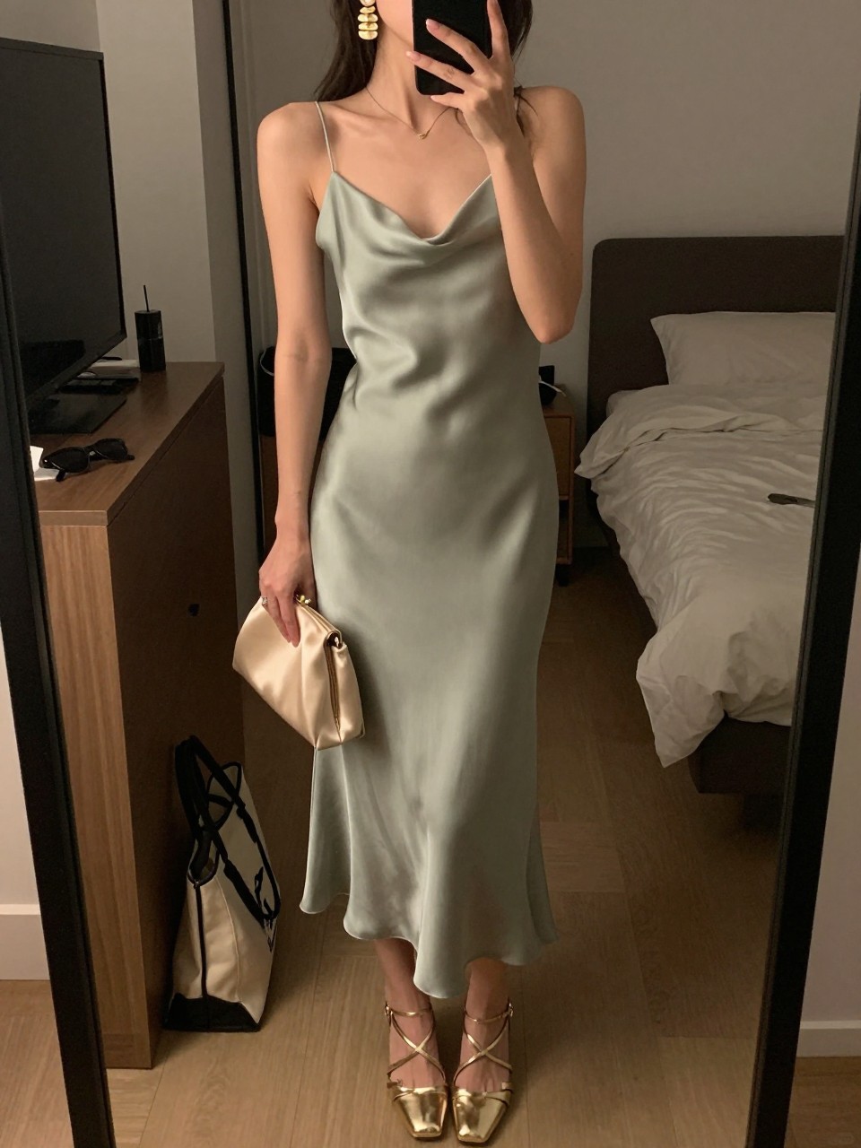 mirror selfie of a woman wearing a sage green silk slip dress, gold statement drop earrings, metallic gold strappy heels, and a small champagne satin clutch, holding a phone in front of a mirror, cropped at NECK so face doesn't show, with a natural casual selfie pose, in a city apartment, warm moody lighting, including small lived-in details like a tote bag on the floor, sunglasses on a dresser, or soft bedding in the background, latest iPhone photo quality.