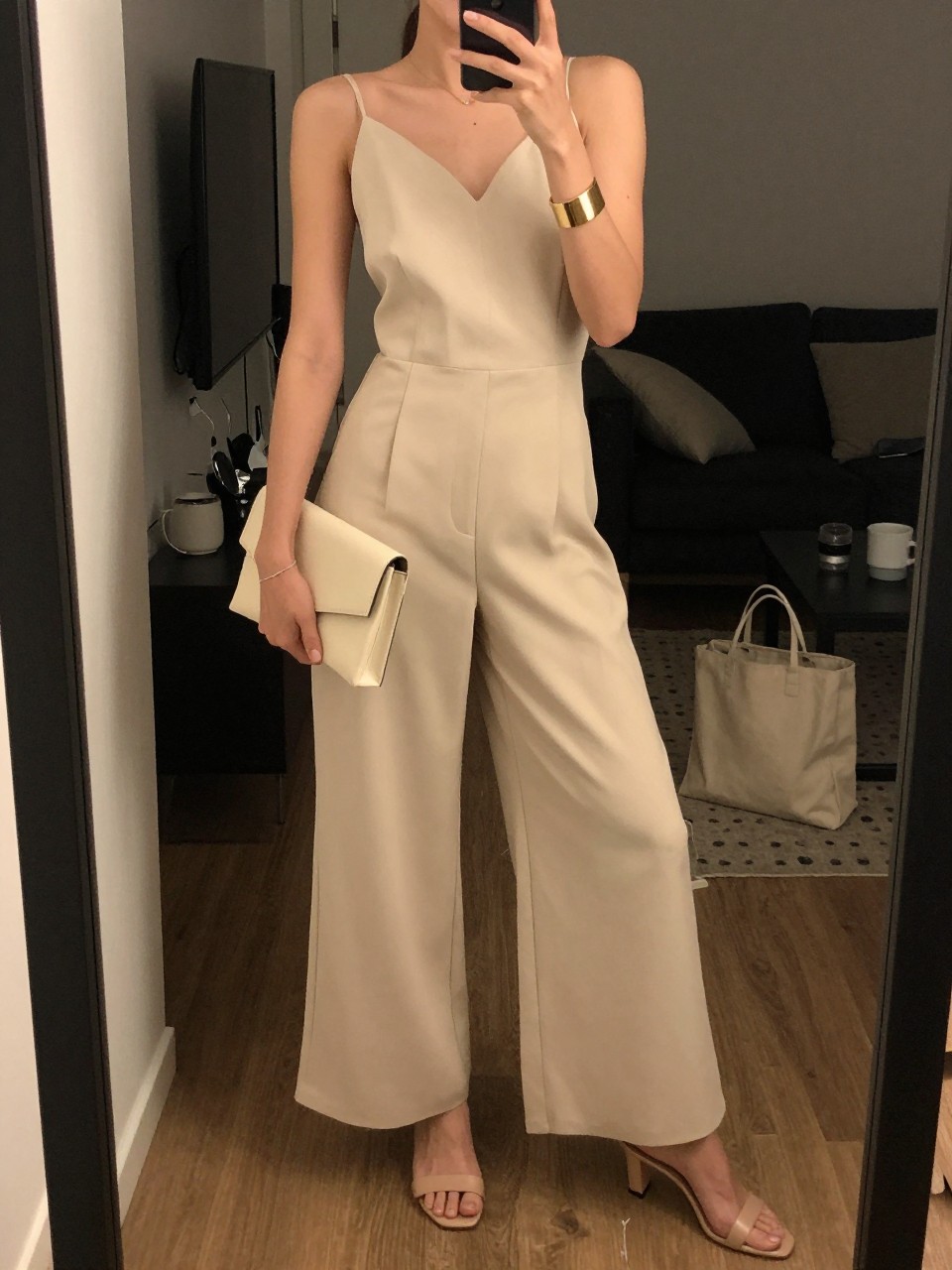 mirror selfie of a woman wearing a champagne wide-leg jumpsuit with nude heeled mules, an ivory clutch, and a gold cuff bracelet, holding a phone in front of a mirror, cropped at NECK so face doesn't show, with a natural casual selfie pose, in a city apartment, warm moody lighting, including small lived-in details like a tote bag on the floor and a coffee mug nearby, latest iPhone photo quality.