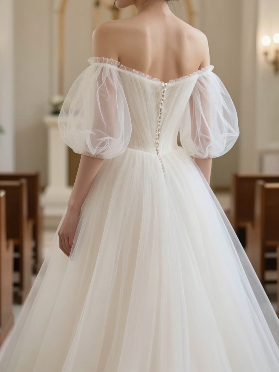 Image Prompt: A dreamy bridal editorial photo of a romantic off-shoulder wedding gown styled with a soft tulle bolero. Show the bride from the shoulders down or from a back view with the face hidden. The bolero is sheer and airy with delicate gathered sleeves, layered over a softly draped gown. Set the image in a bright chapel or elegant indoor venue with gentle light, realistic tulle movement, luxury bridal editorial styling, no visible face, no text, no watermark. (no watermarks on images)