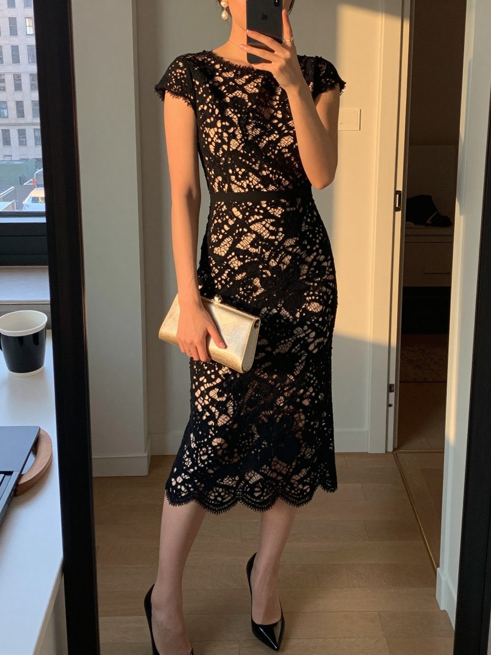 mirror selfie of a woman wearing a black lace midi dress with sleek heels, pearl drop earrings, and a champagne clutch, holding a phone in front of a mirror, cropped at NECK so face doesn't show, with a natural casual selfie pose, in a city apartment, evening golden hour, including small lived-in details like a coffee mug nearby and an open closet behind, latest iPhone photo quality.