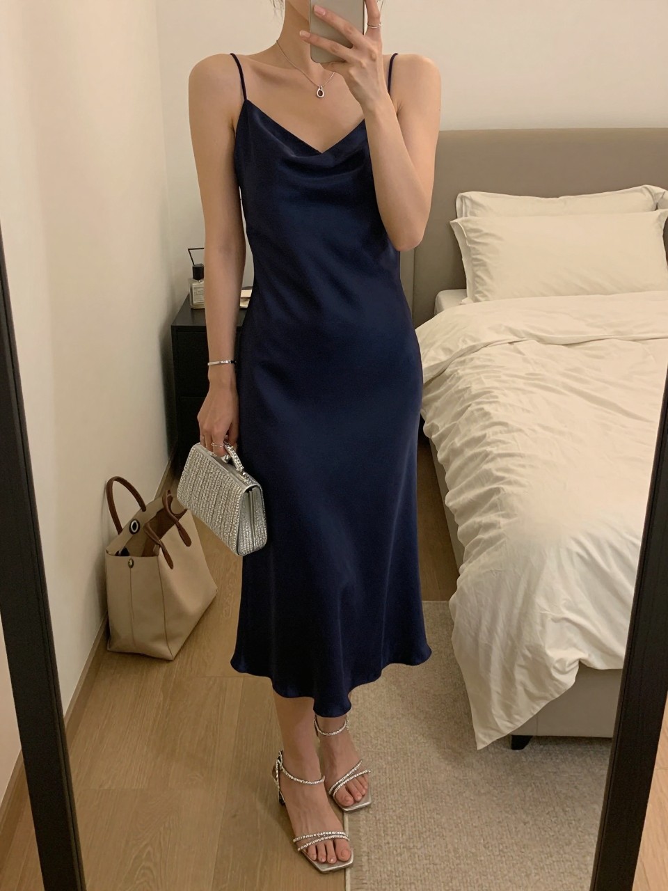 mirror selfie of a woman wearing a navy satin midi slip dress with crystal strappy sandals, a silver clutch, and silver jewelry, holding a phone in front of a mirror, cropped at NECK so face doesn't show, with a natural casual selfie pose, in a feminine-themed bedroom, warm moody lighting, including small lived-in details like a tote bag on the floor and soft bedding in the background, latest iPhone photo quality.