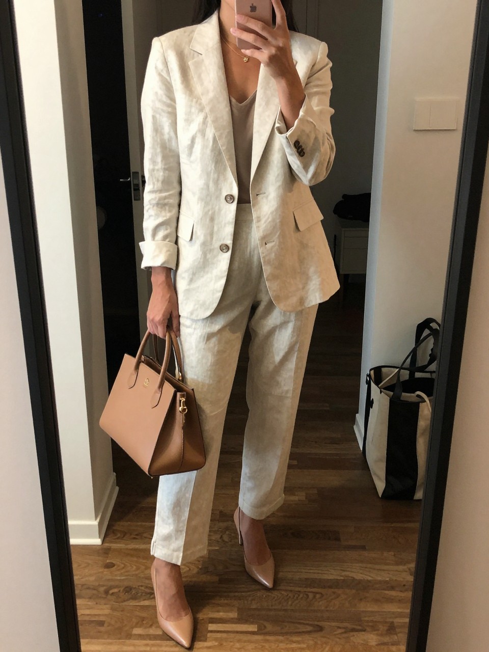 mirror selfie of a brown woman wearing a cream linen blazer with matching tailored linen trousers, nude pointed-toe heels, a tan structured bag, and a slim gold necklace, holding a phone in front of a mirror, cropped at NECK so face doesn't show, with a natural casual selfie pose, in a city apartment, warm moody lighting, including small lived-in details like an open closet behind and a tote bag on the floor, latest iPhone photo quality.