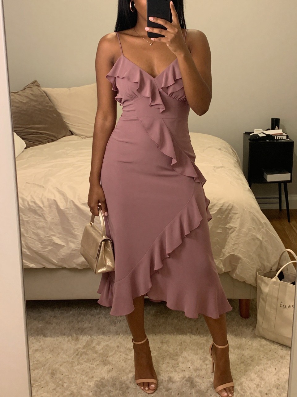mirror selfie of a black woman wearing a mauve ruffle chiffon midi dress with nude ankle-strap heels, a champagne mini bag, and rose gold earrings, holding a phone in front of a mirror, cropped at NECK so face doesn't show, with a natural casual selfie pose, in a feminine-themed bedroom, warm moody lighting, including small lived-in details like soft bedding and a tote bag on the floor, latest iPhone photo quality.