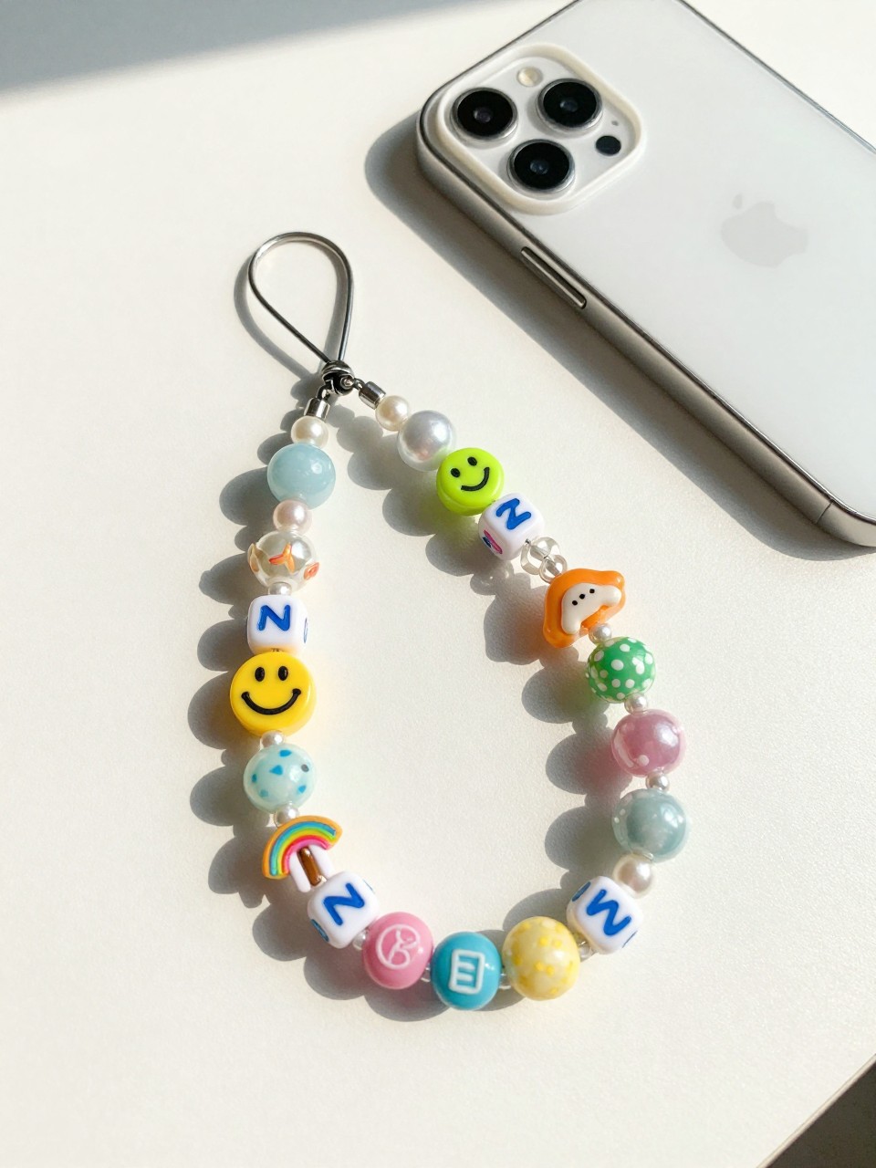 Detailed Image Prompt:
A stylish editorial craft photo of handmade beaded phone charms for teens, colorful glass beads, letter beads, smiley face beads, pearl beads, and small summer charms arranged on a clean white desk, finished phone charms clipped to a phone case, soft natural daylight, trendy teen craft aesthetic, bright summer colors, no hands, no faces, no readable text, no watermark on images.