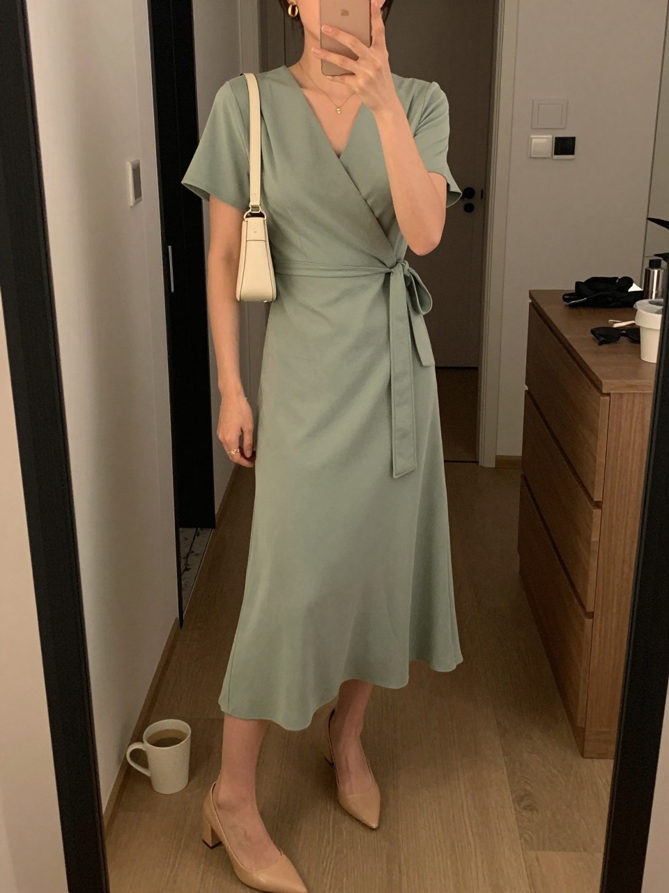 mirror selfie of a woman wearing a sage green wrap dress with nude block heels, gold hoop earrings, and a cream mini shoulder bag, holding a phone in front of a mirror, cropped at NECK so face doesn't show, with a natural casual selfie pose, in a city apartment, warm moody lighting, including small lived-in details like a coffee mug nearby, an open closet behind, or sunglasses on a dresser, latest iPhone photo quality.