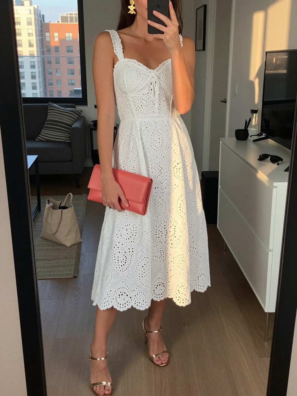 mirror selfie of a woman wearing a white eyelet midi dress with metallic champagne heeled sandals, gold statement earrings, and a coral clutch, holding a phone in front of a mirror, cropped at NECK so face doesn't show, with a natural casual selfie pose, in a city apartment, evening golden hour, including small lived-in details like a tote bag on the floor and sunglasses on a dresser, latest iPhone photo quality.