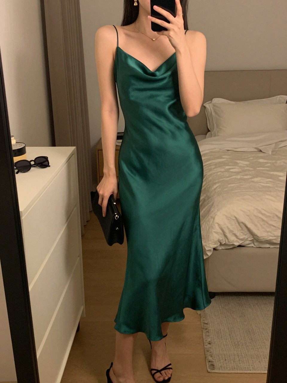 mirror selfie of a woman wearing an emerald green satin midi dress with black strappy heels, a small black clutch, and gold drop earrings, holding a phone in front of a mirror, cropped at NECK so face doesn't show, with a natural casual selfie pose, in a feminine-themed bedroom, warm moody lighting, including small lived-in details like sunglasses on a dresser and soft bedding in the background, latest iPhone photo quality.