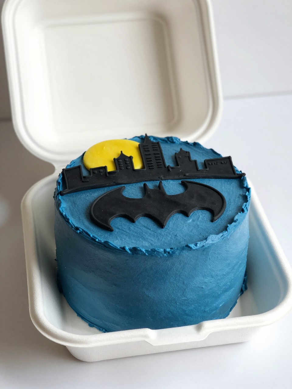 Image Prompt:
A Batman themed bento cake in an open white cake box, smooth deep blue frosting base, black piped Gotham city skyline around the top edge, yellow moon detail with a small bat symbol, neat modern superhero styling, realistic editorial dessert photography, soft natural light, no people, no hands, no faces, (no watermarks on images)