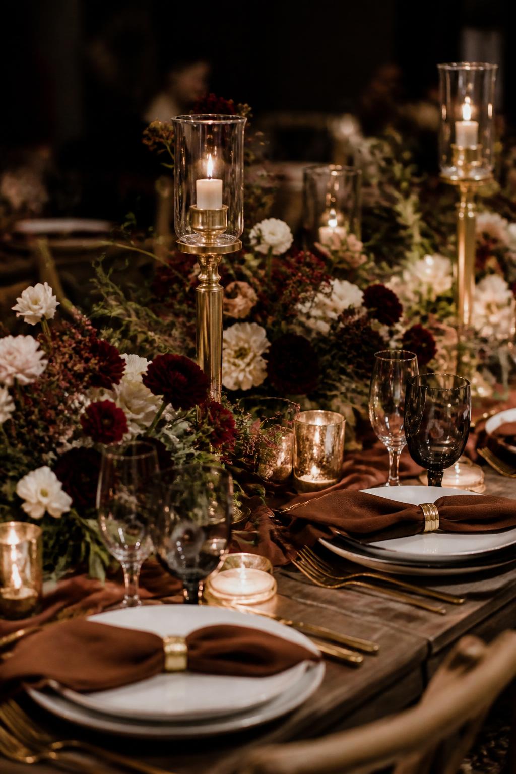 Image Prompt:
A dramatic fall wedding table styled with brown linen napkins, burgundy flowers, ivory blooms, dark greenery, gold candle holders, white plates, smoky glassware, and warm low lighting. Elegant evening reception setting, no visible faces, no text, no watermark, high-end DSLR wedding photography, (no watermarks on images)