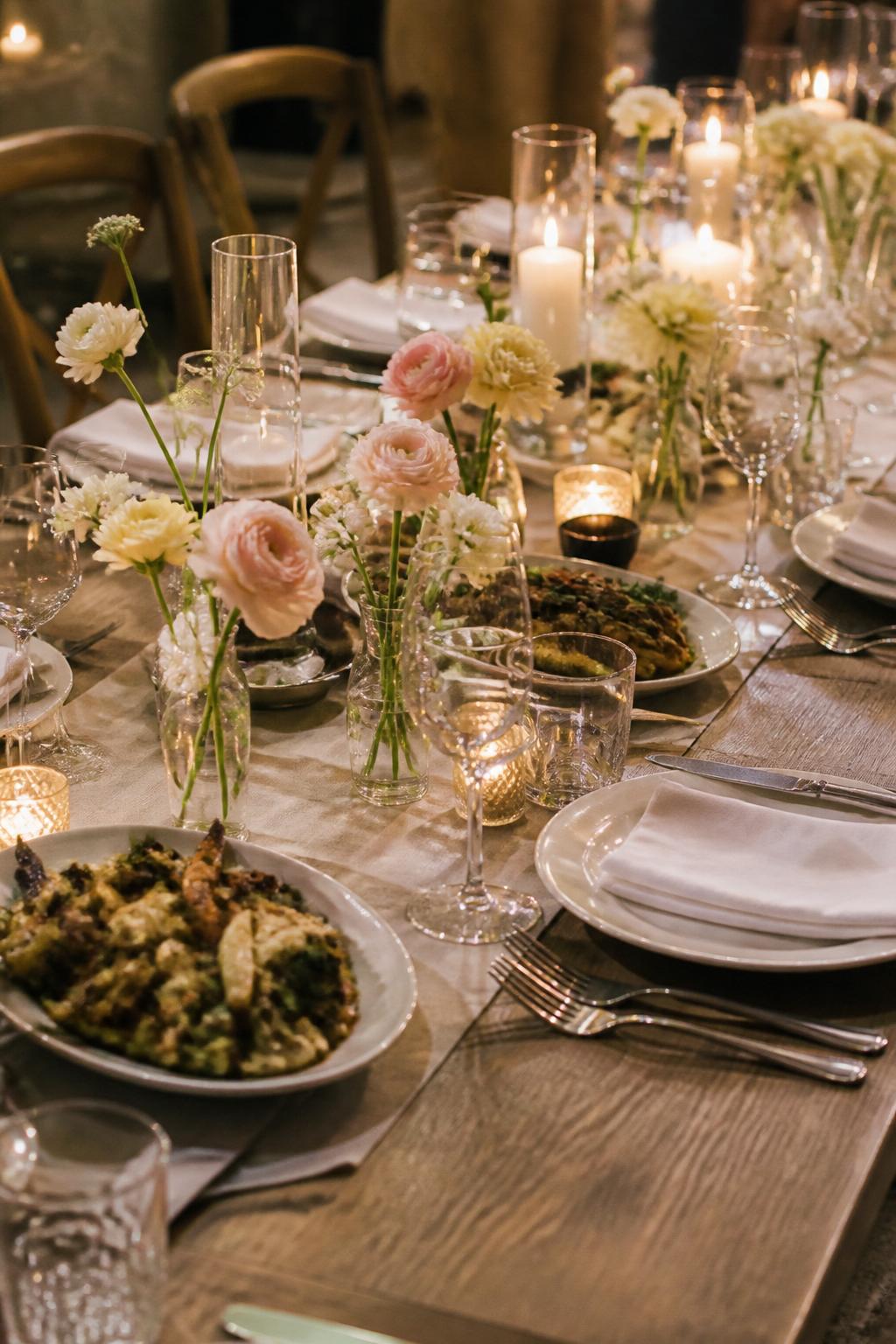Image Prompt:
A beautiful family style wedding dinner table with small clear bud vases placed between shared serving platters, each vase holding one or two flowers in white, blush, and pale yellow. Add white plates, linen napkins, clear glasses, polished flatware, and soft candlelight. Intimate reception setting, no visible faces, no text, no watermark, realistic editorial wedding photography, (no watermarks on images)