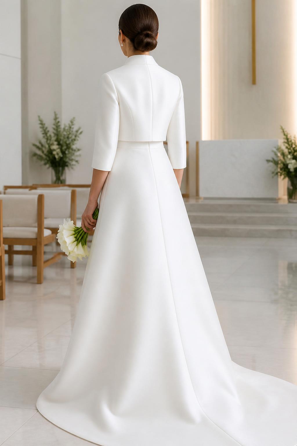 Image Prompt: A professional bridal editorial photo of a minimalist A-line wedding dress paired with a structured mikado bolero. Show the bride from the shoulders down or in a back-facing pose so the face is not visible. The dress has a clean silhouette and the bolero features a crisp tailored shape, modest neckline, and elegant three-quarter sleeves. Place the scene in a bright modern chapel or luxury bridal studio with clean lines, realistic mikado texture, magazine-quality photography, no visible face, no text, no watermark. (no watermarks on images)