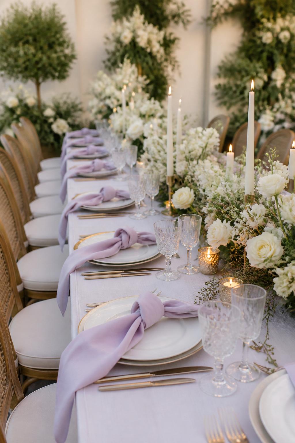 Image Prompt:
A professional editorial wedding reception photo showing a long rectangle wedding table styled with lavender linen napkins, low white floral centerpieces, soft greenery, ivory taper candles, white plates, gold flatware, and clear crystal glasses. Elegant garden or indoor wedding reception setting, no visible faces, no full people, table-focused composition, realistic DSLR wedding photography, no text, no watermark, (no watermarks on images)