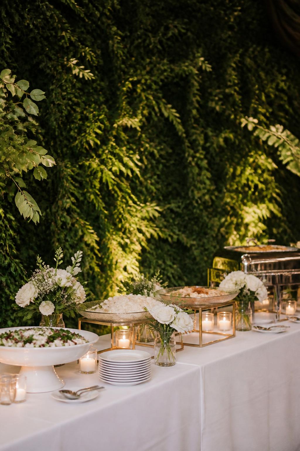 Image Prompt:
A stylish wedding buffet table placed in front of a greenery wall or lush greenery backdrop. The table is styled with white linen, elegant serving dishes, small floral arrangements, clear glass bowls, white plates, and warm soft lighting. No visible faces, no full people, buffet table-focused shot, no text, no watermark, realistic editorial wedding photography.