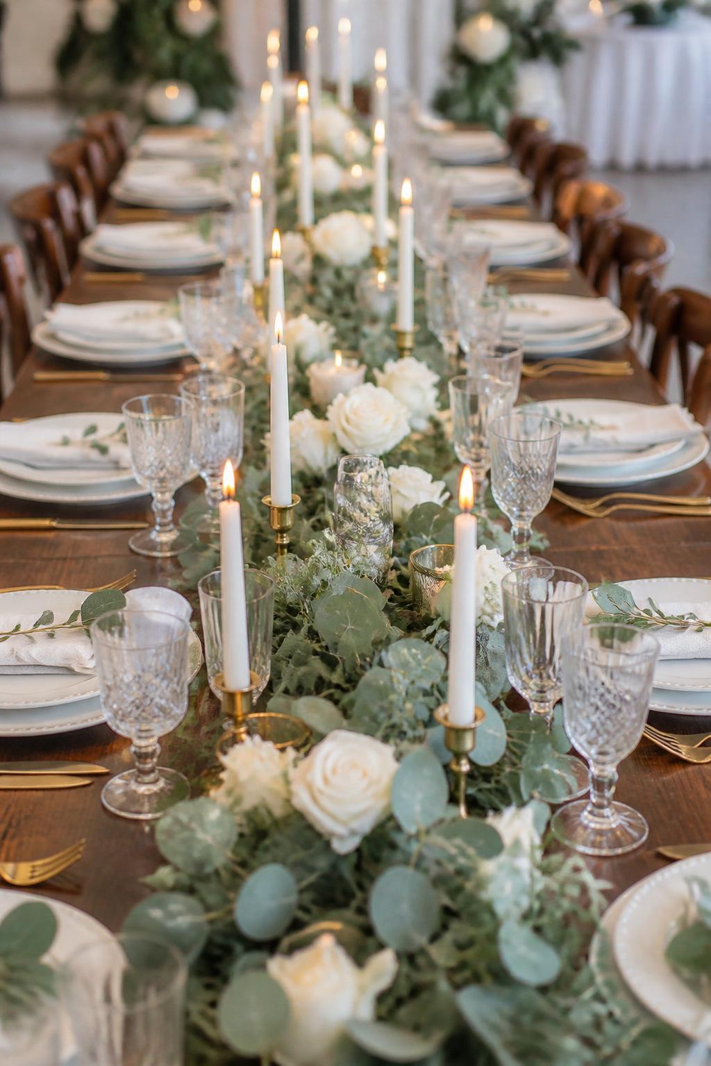 Image Prompt:
A professional editorial wedding reception photo showing a long rectangle wedding table styled with a lush eucalyptus garland running down the center, white roses, ivory taper candles in elegant holders, white plates, folded linen napkins, clear crystal glasses, and polished gold or silver flatware. Elegant garden or indoor wedding reception setting, no visible faces, no full people, table-focused composition, realistic DSLR wedding photography, no text, no watermark, (no watermarks on images)