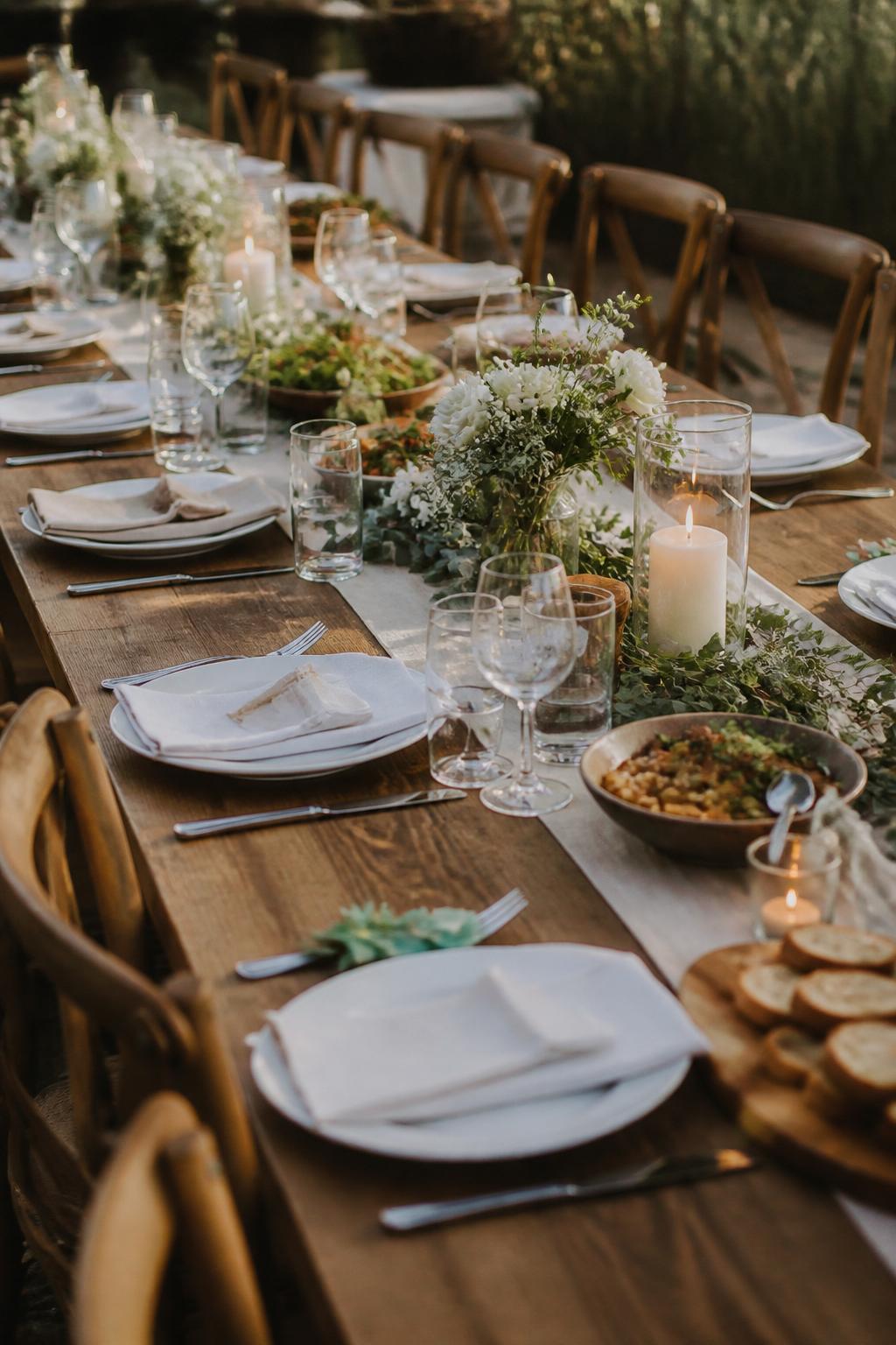 Image Prompt:
A warm family style wedding dinner table on a long wooden table, styled with simple white plates, folded linen napkins, clear glassware, low greenery, small candles, shared ceramic platters, and soft white floral touches. Barn, backyard, or vineyard reception setting, no visible faces, no full people, no text, no watermark, realistic wedding photography, (no watermarks on images)