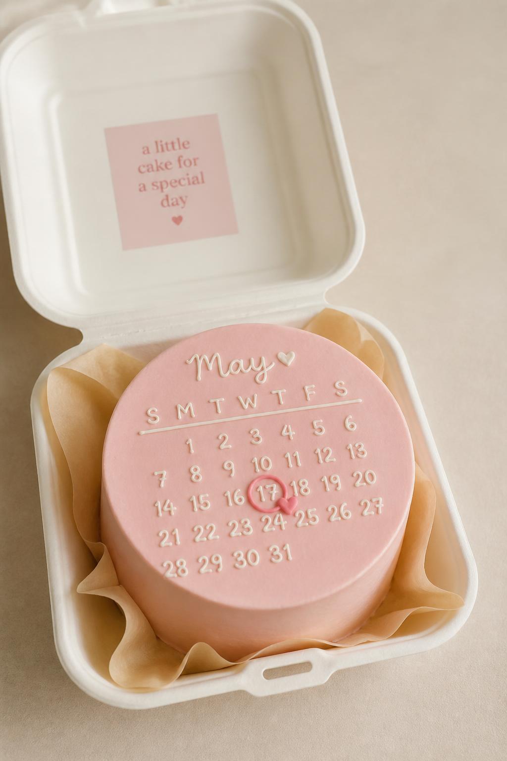 A personalized pink bento cake designed like a tiny calendar page, smooth pale pink frosting, thin white icing calendar lines, one date circled in darker pink with a tiny heart, open white bento cake box, clean editorial styling, soft beige background, realistic bakery photography, no people, no hands, no faces, no watermarks on images