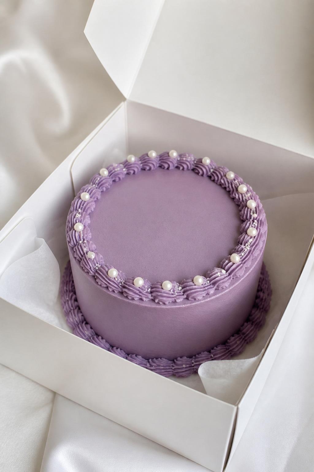 Image Prompt:
A beautiful purple pearl bento cake inside a white cake box, smooth dusty purple frosting, tiny edible pearl accents placed neatly around the top edge, elegant piped border, soft luxury bakery styling, clean backdrop, realistic editorial dessert photography, no people, no hands, no faces, (no watermarks on images)