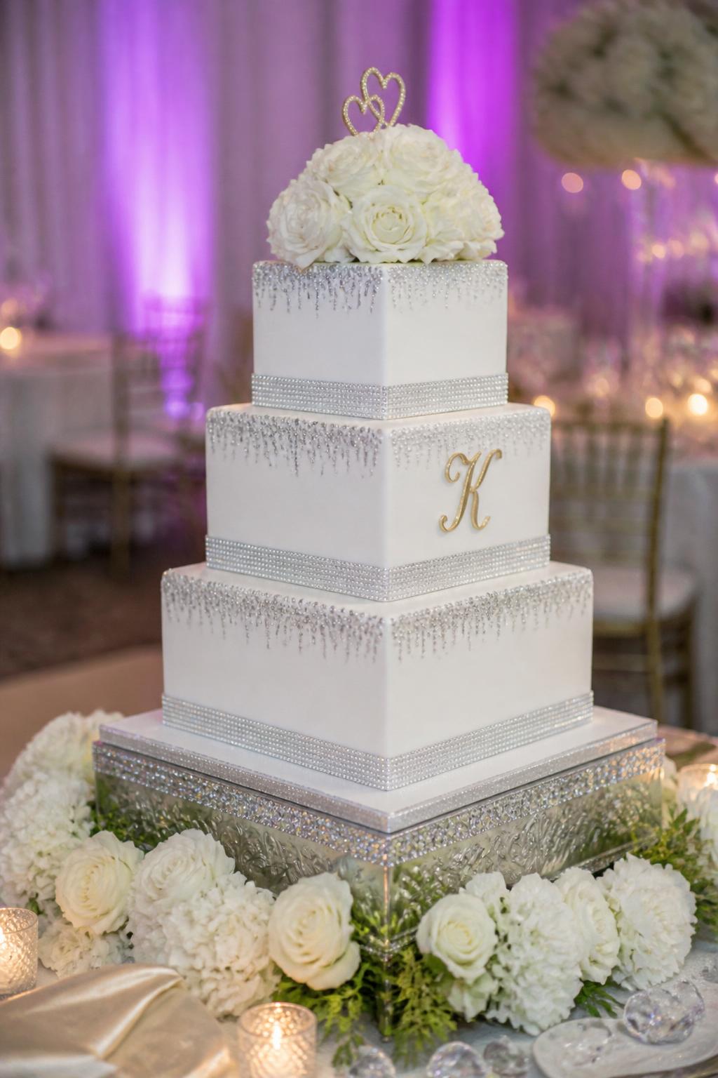 Image Prompt: Ultra-realistic modern wedding reception photo of a square tier bling wedding cake with sharp clean edges, crystal bands, and refined metallic accents. Style the cake at a real wedding party scene with modern decor, floral arrangements, candles, and elegant reception details. The composition should look like a real professional wedding photo with the cake as the clear main focus. No faces visible. If any people appear, show only blurred side views, hands, or back views. Realistic textures, beautiful light, and a modern upscale feel. (no watermarks on images)